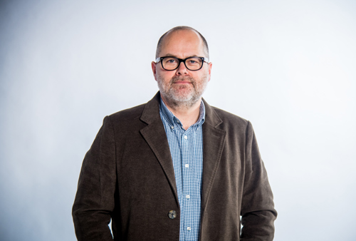 Mark our Chief Exec stands smiling at the camera. He is wearing glasses and a brown jacket with a blue shirt.